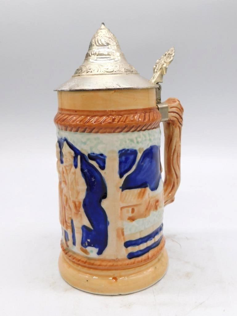 German-Style Ceramic Stein Decanter III: A ceramic stein-form whiskey decanter, one of a small group in this collection, its body worked in cobalt blue with a relief village scene. The handle is finished in wood grain and the lid is hinged
