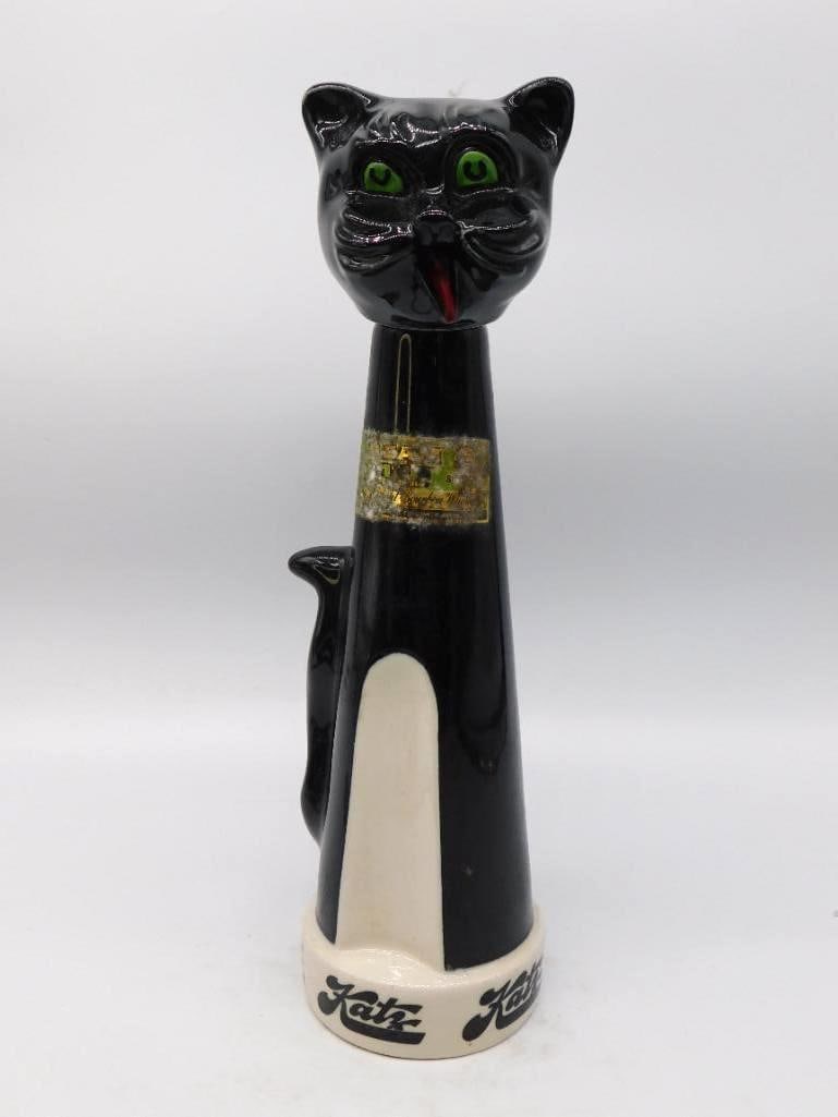 Katz Black Cat Decanter, Green-Eyed: A tall ceramic decanter modeled as a long-necked cat, glazed a glossy jet with green eyes and a red tongue, its white breast running down to a base lettered Katz. A jaunty piece of advertising ware