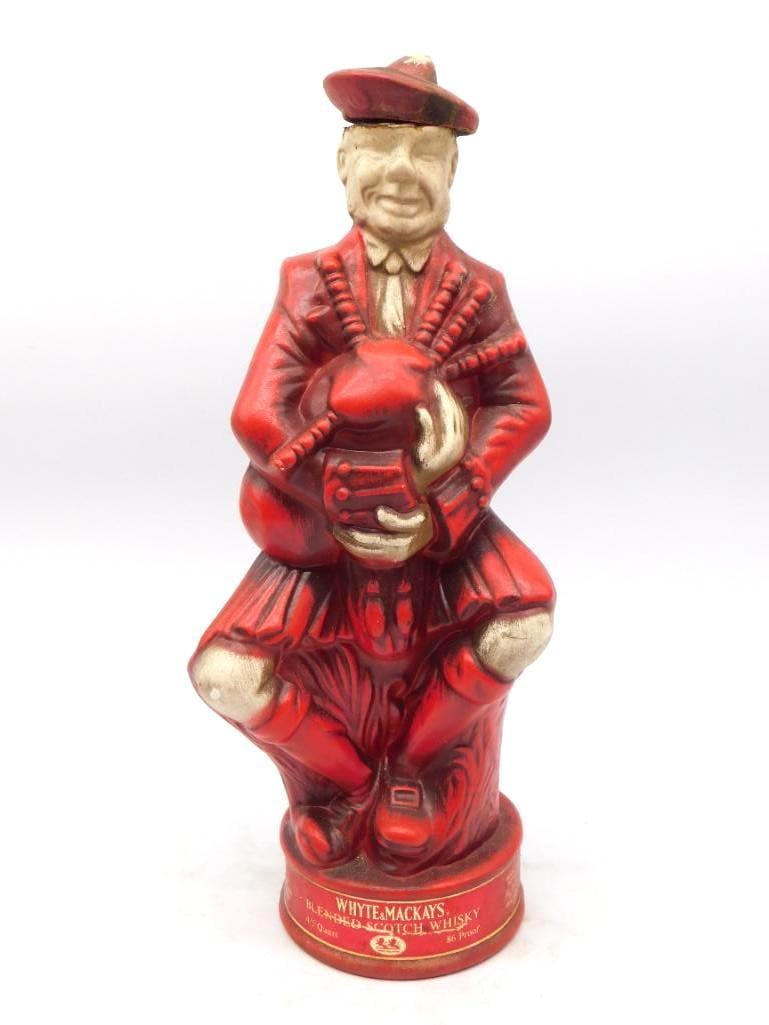 Whyte and Mackays Scottish Piper: A genial decanter modeled as a Scottish piper seated on a stump, bagpipes under his arm and bonnet cocked over a cheerful face, finished in a deep red glaze with cream flesh tones. Approximately 11