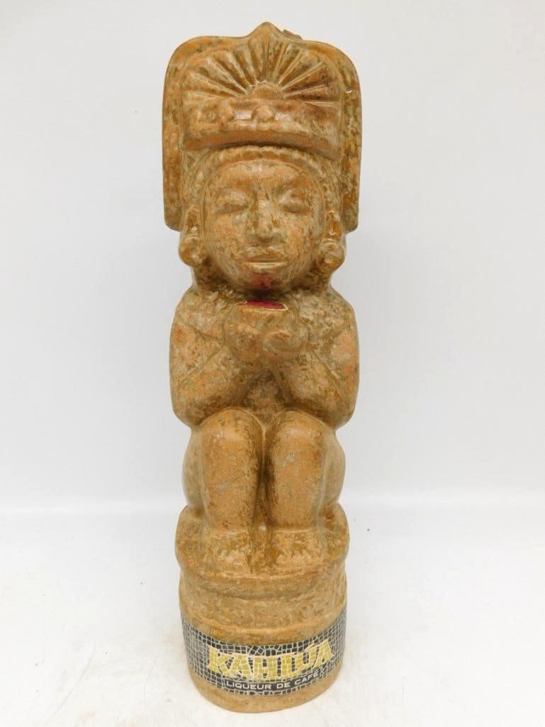 Kahlua Seated Effigy Ceramic Liqueur: A ceramic Kahlua bottle modeled after a seated stone effigy, arms folded at the chest beneath a broad plumed headdress, finished in a mottled stone-color glaze that mimics carved rock. A singular form