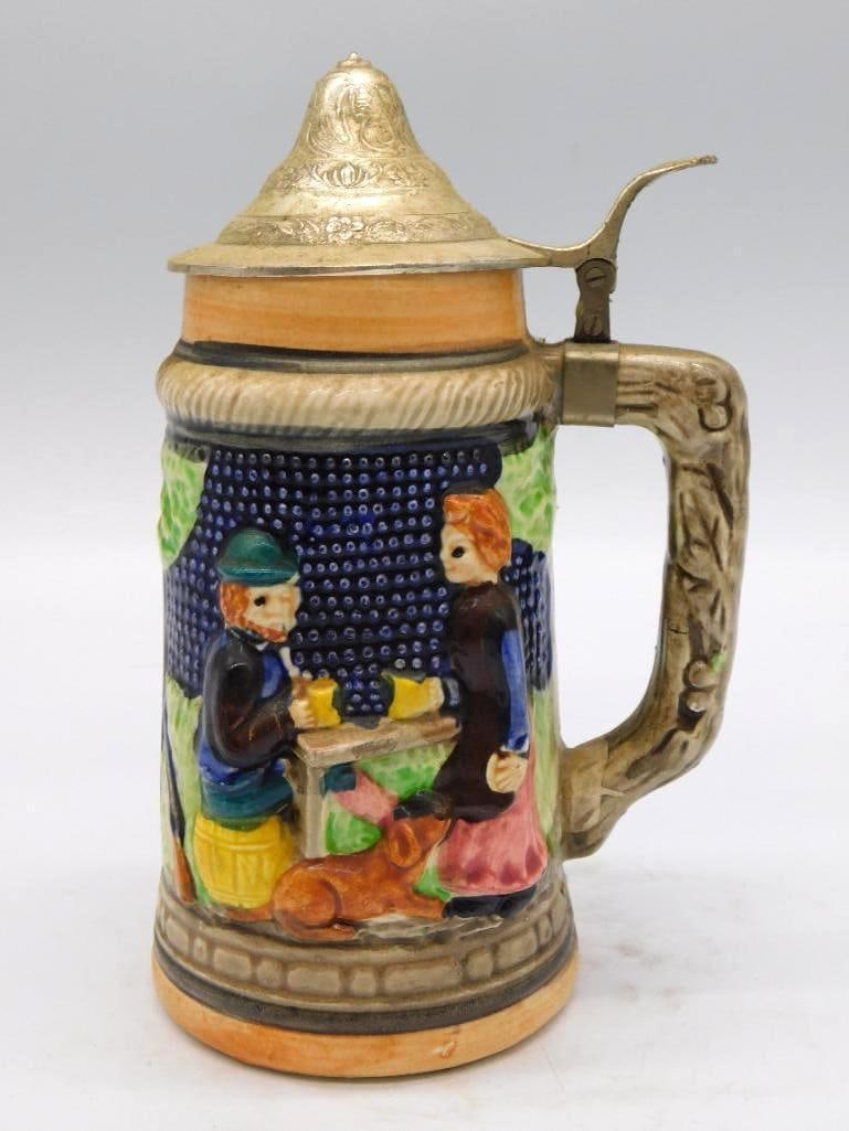 German-Style Majolica Castle Scene: A hand-painted ceramic stein-form decanter with hinged pewter-tone thumb-lift lid, the body relief-molded with a blue castle and village scene, figures set against a green landscape, and an applied go