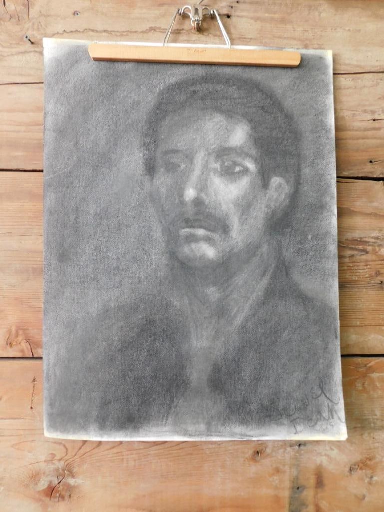 Charcoal Portrait of a Man on Paper: Charcoal portrait depicting a man's face on a textured paper. The artwork is mounted on a board with a clip at the top for support. The artist's signature appears faintly on the bottom corner. A