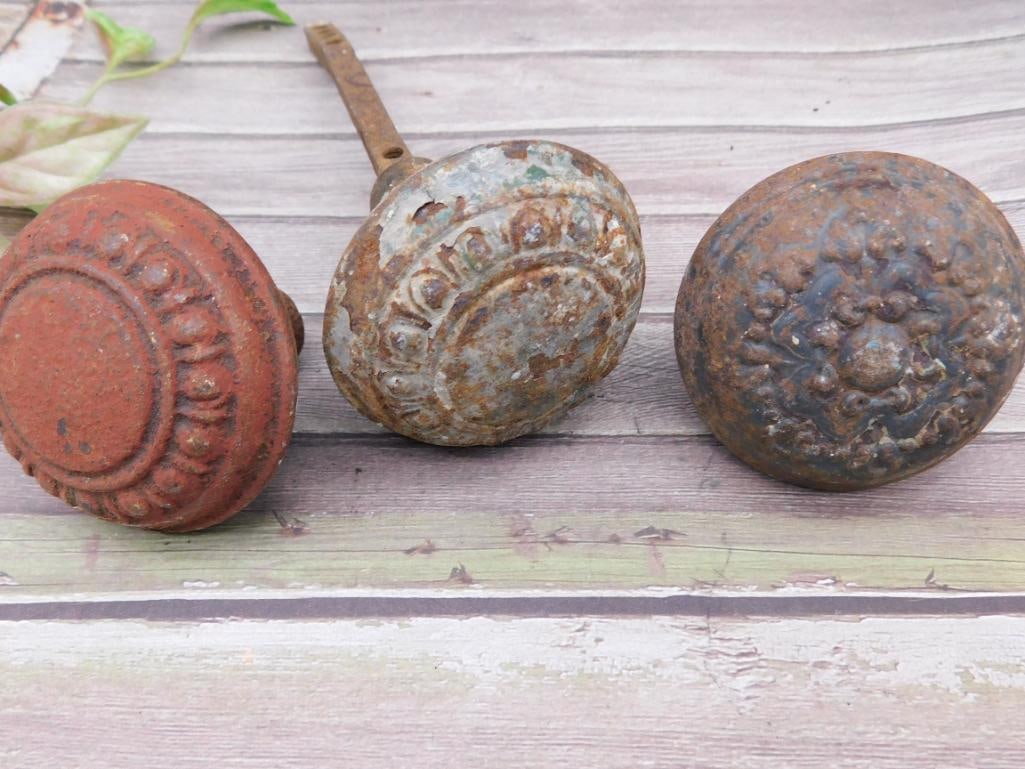 Set of Three Cast Iron Door Knobs with Decorative Patterns: This lot includes three decorative cast iron door knobs. Each knob displays varying patinas and levels of rust, highlighting their aged condition. One knob includes a metal spindle attachment. These