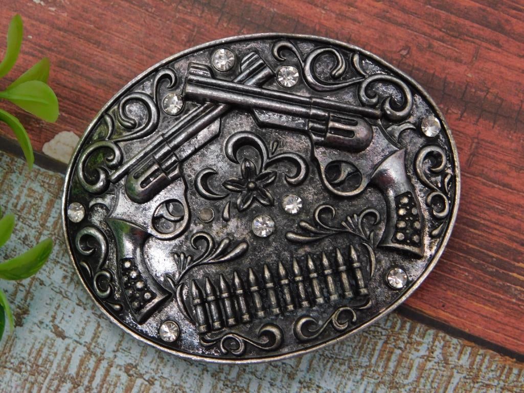 Western Belt Buckle with Crossed Pistols and Rhinestones: Oval metal belt buckle featuring an embossed design of crossed revolvers, a bullet band, and floral scrollwork, accented with clear rhinestones. Antiqued silver-tone finish shows expected wear.