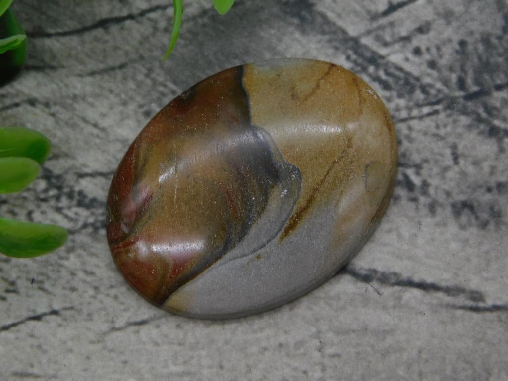 Rock: Polished Picture Jasper Palm Stone Cabochon: Polished oval jasper palm stone displaying banded red, tan, and gray coloring reminiscent of a landscape scene. Measures approximately 2.25 inches long (quarter shown for scale), with a smooth polishe