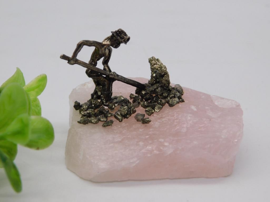 Figurine - Miner on Rose Quartz Base with Pyrite Ore Clusters: Miniature bronze-tone figural sculpture of a laborer swinging a pickaxe, mounted atop a natural rose quartz stone base studded with clusters of sparkling pyrite crystals arranged to resemble an ore ve