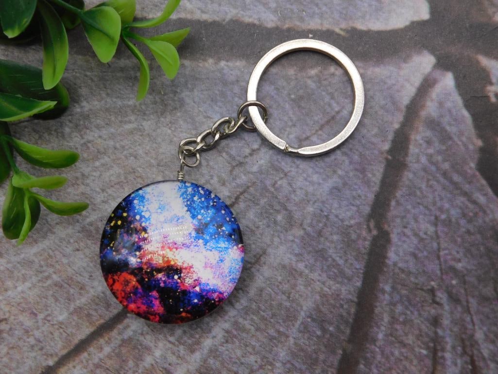 Keychain Galaxy Resin Pendant Cosmic Design: Round resin pendant keychain featuring a hand-poured "galaxy" design in swirling blue, red, and white cosmic colors with glitter accents. Pendant measures approximately 1.5 inches in diameter, shown