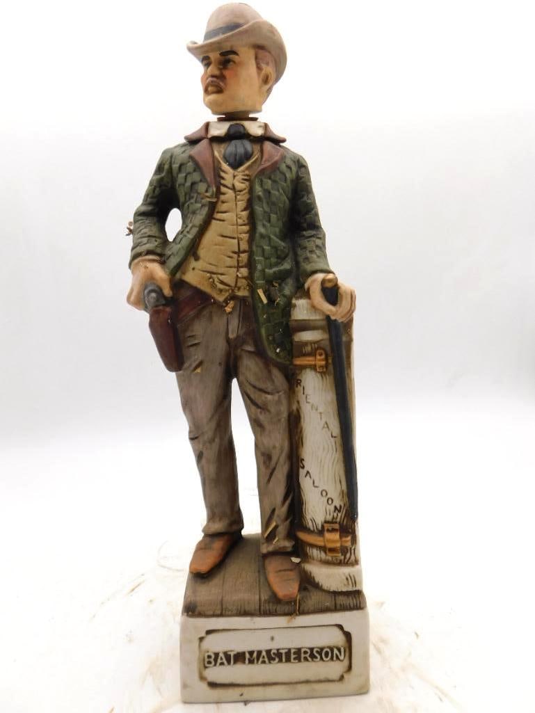 Bat Masterson Old West Lawman Decanter: A ceramic decanter modeled as the Western lawman Bat Masterson, bottle in hand beside an "Oriental Saloon" prop of golf-bag form, the base marked "Bat Masterson." A storied name of the frontier,