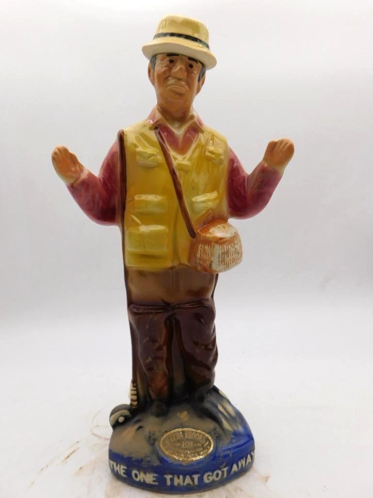 Ezra Brooks The One That Got Away Fisherman Ceramic Decanter: An Ezra Brooks ceramic decanter modeled as a smiling fisherman in tackle vest and straw hat telling the tale with his hands. The base is inscribed The One That Got Away and the original Ezra Brooks