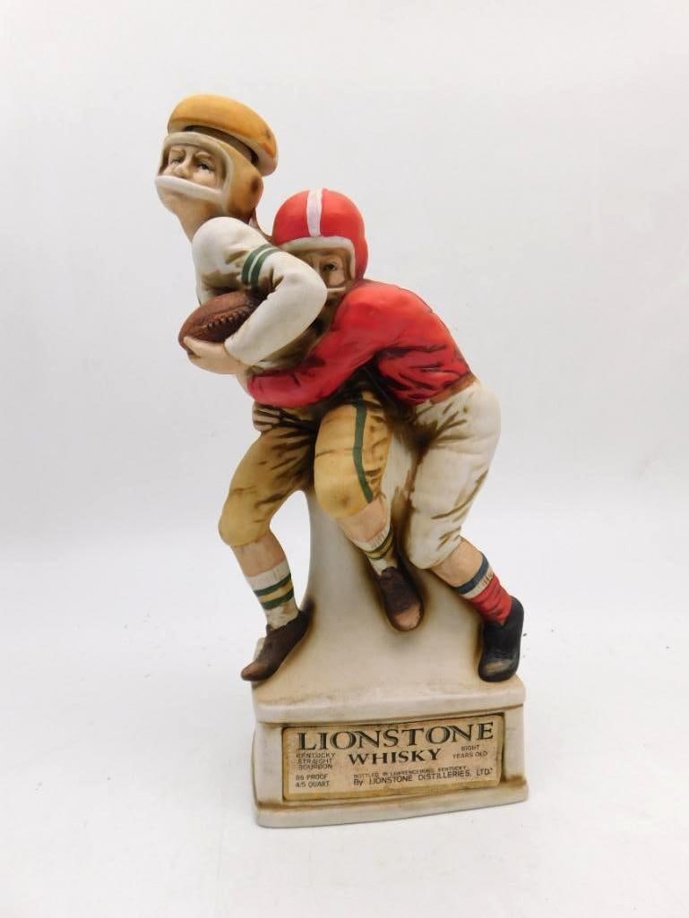 Lionstone Football Players Tackle Whiskey Decanter: A Lionstone porcelain decanter modeled as two players in vintage uniform locked in a tackle one with the ball tucked under his arm upon a plaque base carrying descriptive text on the history of footba
