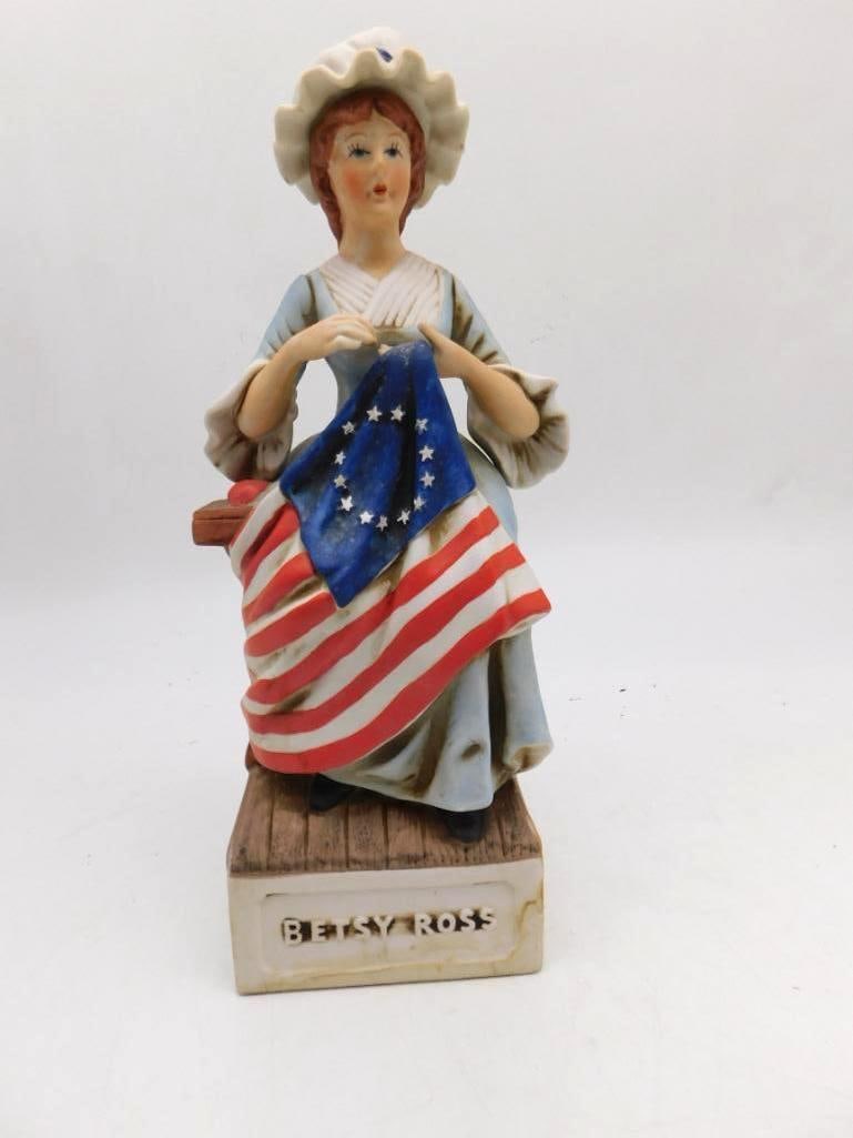 McCormick Betsy Ross Porcelain Whiskey Decanter: A McCormick Distilling Co. porcelain decanter modeled as Betsy Ross seated at her sewing of the American flag. The wooden base is marked Betsy Ross. A limited edition labeled Straight Bourbon