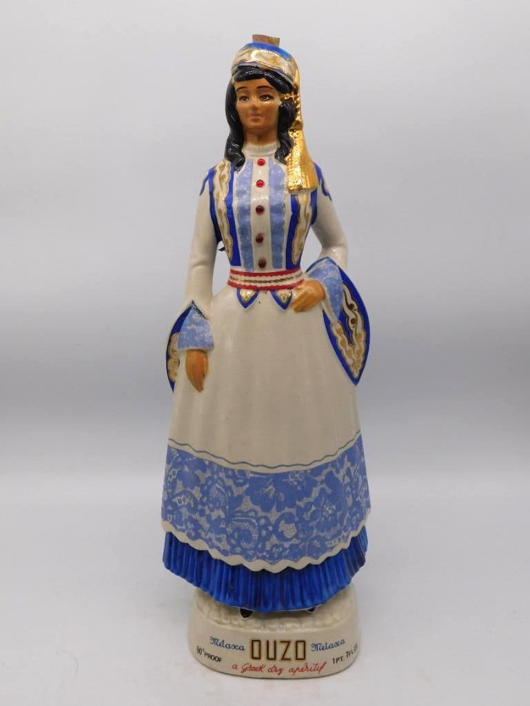 Metaxa Ouzo Greek Maiden Decanter in Traditional Blue and White Dress: A trim porcelain decanter modeled as a Greek woman in traditional dress her gown white and blue with gold braid at the bodice and a red sash at the waist. The painter worked a fine hand on the lacewor