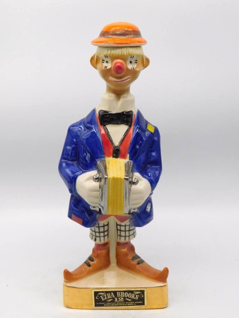 Ezra Brooks Clown with Accordion Whiskey Decanter 12 Years Old: A genial Ezra Brooks decanter modeled as a circus clown in a blue coat and orange straw hat an accordion squeezed between his white gloves. The painted face is cheerful without turning coarse and the 