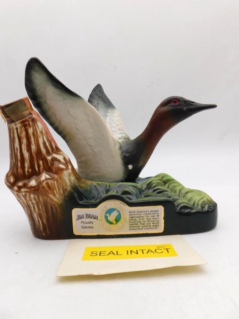 Jim Beam Ducks Unlimited Canvasback Duck Decanter Seal Intact: A Jim Beam decanter modeled as a canvasback drake rising from the marsh wings spread and neck stretched low above sculpted grasses beside a weathered stump. The plumage is shaded with an admirable han