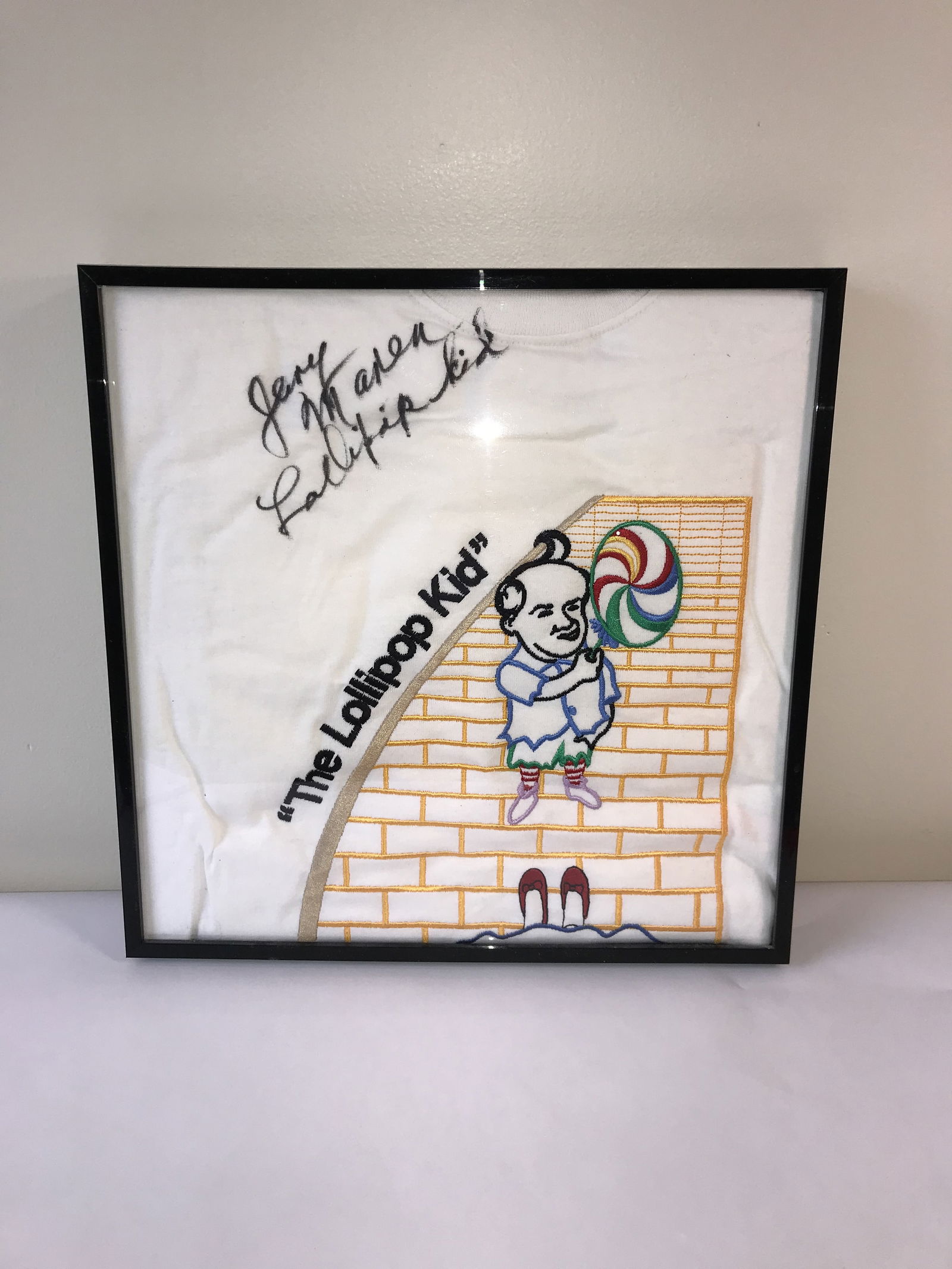 Jerry Maren Signed The Wizard of Oz The Lollipop Kid Framed Embroidered T-Shirt: This lot features a framed white fabric piece, likely a t-shirt, showcasing embroidered graphics dedicated to the 1939 cinematic masterpiece The Wizard of Oz. The embroidery depicts a member of the Lo
