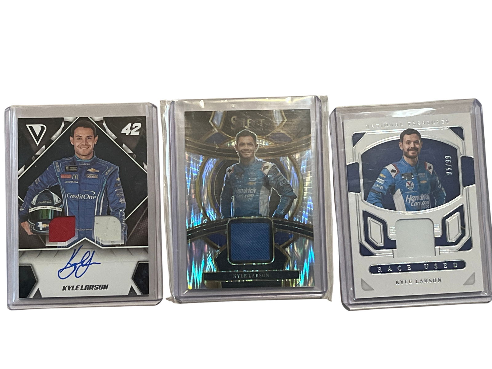 A Trio of Panini Kyle Larson NASCAR Relic Trading Cards Including Autograph: A trio of Panini NASCAR trading cards featuring driver Kyle Larson, issued between 2019 and 2024. The lot includes a 2019 Victory Lane dual relic card with a blue ink autograph (DSS-KL), a 2023 Nation