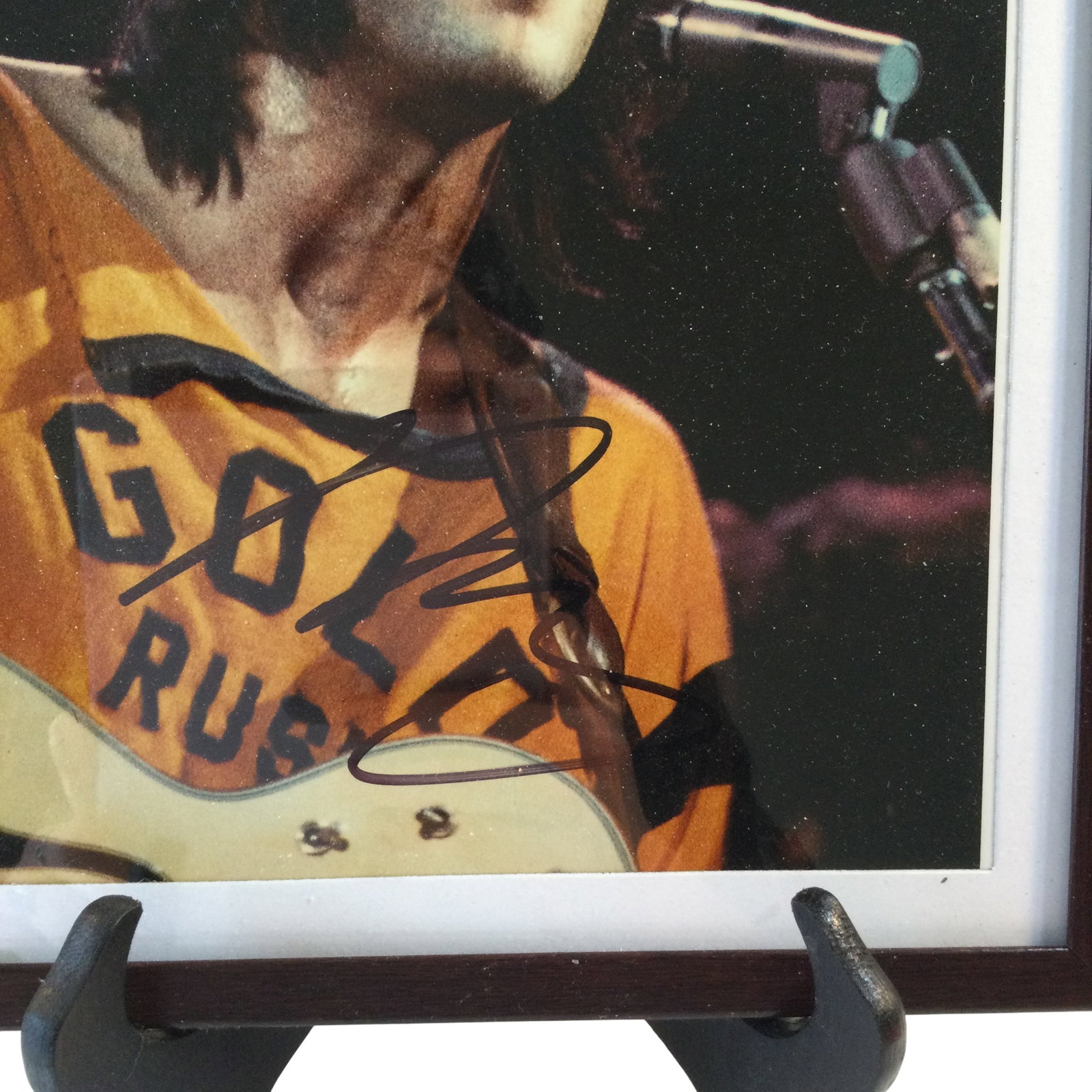 Neil Young Framed Autographed Photograph: A framed color photograph of musician Neil Young depicted during a live performance. The subject is shown from the chest up, singing into a microphone while playing a guitar. He is wearing an orange t