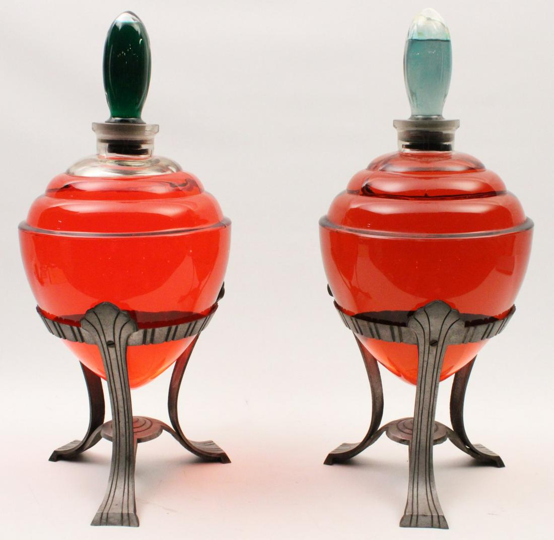 PR. OF CAPPED GLASS PHARMACY GLOBES ON STANDS: PAIR OF CAPPED GLASS PHARMACY GLOBES WITH ART DECO INFLUENCED METAL TRIFORM STANDS, MID 20TH C.; 23"H X 10"W OVERALL; CONDITION: 1 CAPPED GLOBE DAMAGED AND RE-GLUED