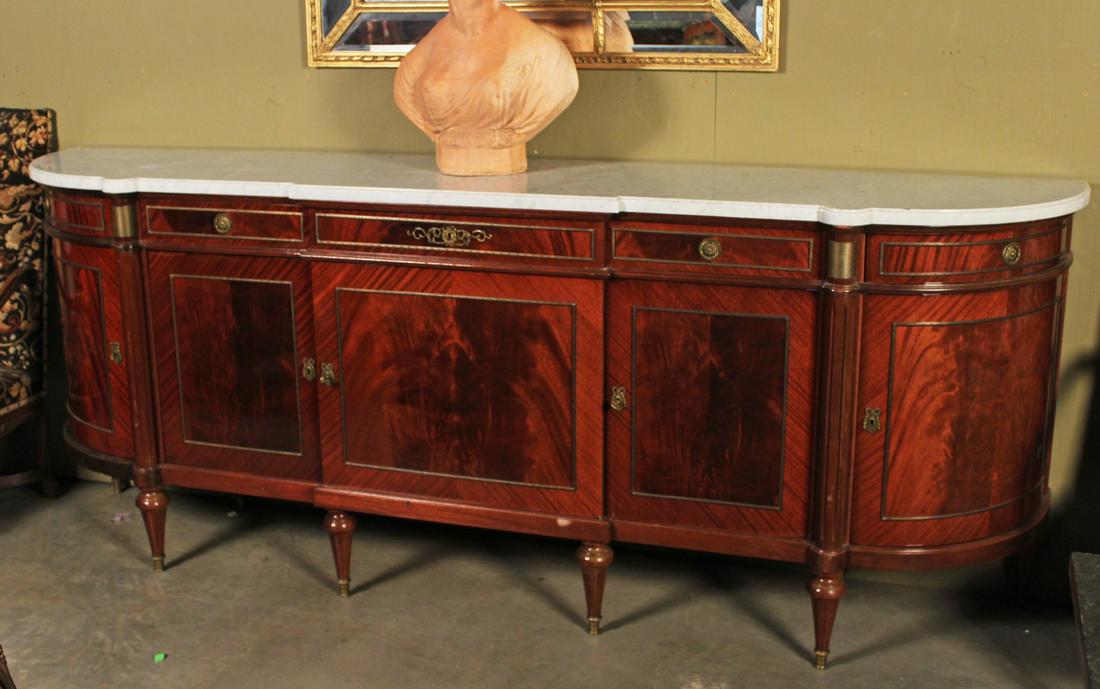 LOUIS XVI STYLE CROTCH MAHOGANY GRAND BUFFET: LOUIS XVI STYLE CROTCH MAHOGANY STEPFRONT GRAND BUFFET HAVING WHITE AND GREY VEINED CARRARA MARBLE TOP OVER 3 DRAWERS AND 2 RADIUS DRAWERS, THEN 5 CONFORMING CUPBOARD DOORS, SUPPORTED BY BRONZE CAPITA