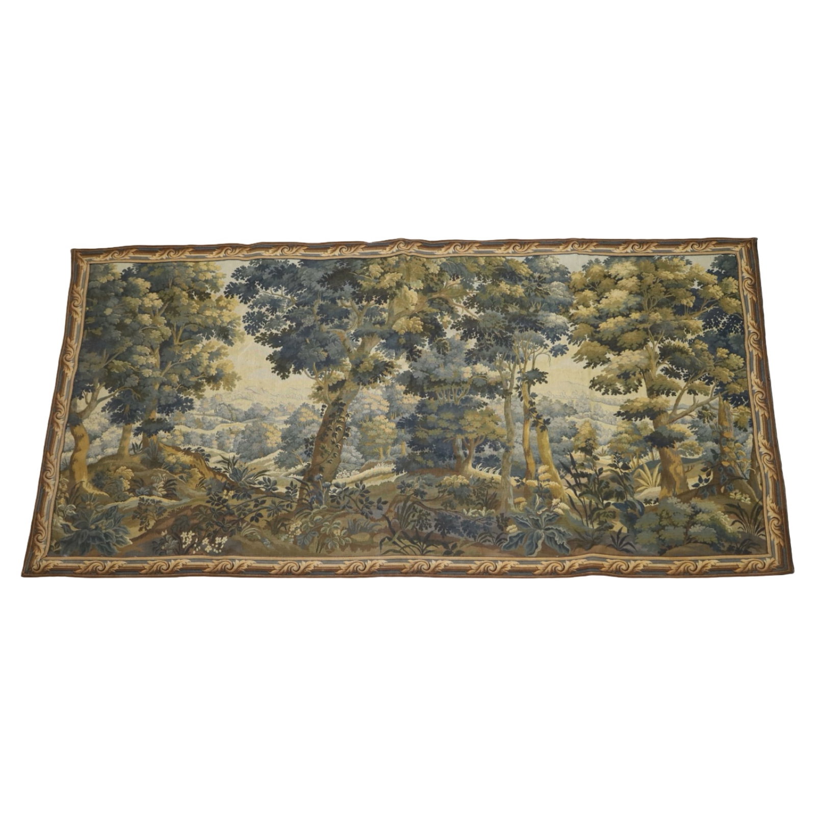 EUROPEAN VERDURE TAPESTRY: Decorative European verdure style woven tapestry depicting an expansive wooded landscape with distant hills in a palette of blue, olive, and ochre tones. Finished with a decorative woven border and