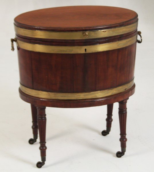 BRASS BOUND OVAL MAHOGANY SHERATON CELLARETTE: BRASS BOUND OVAL MAHONGAY SHERATON CELLARETTE HAVING A HINGED CROSSBANDED TOP OPENING TO AN OPEN WELL ON A TURNED LEG STAND WITH BRASS CASTERS, C. 1820; 27.75"H X 23"W X 17"D