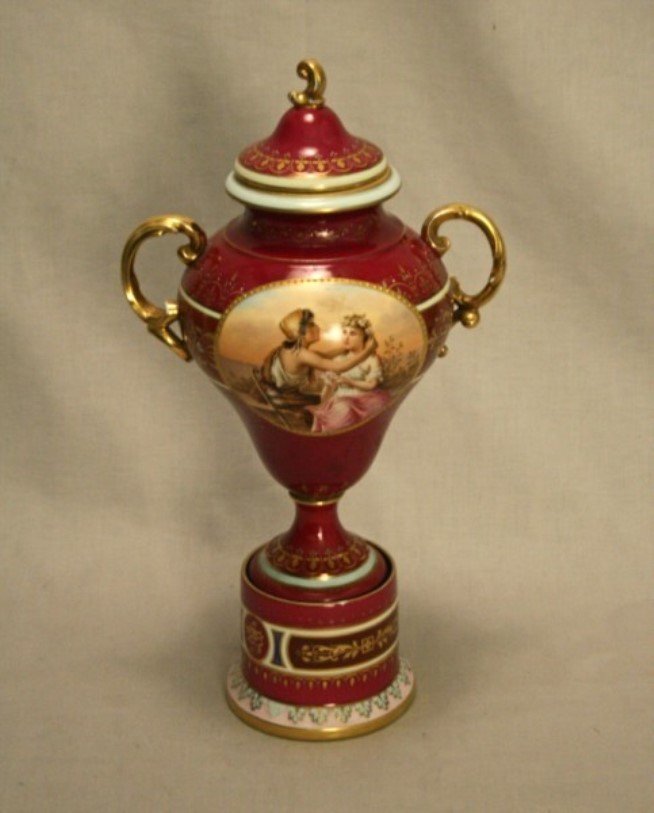 ROYAL VIENNA 15" CAPPED PORCELAIN URN ON CONFORMING: ROYAL VIENNA 15" CAPPED PORCELAIN URN ON CONFORMING PLINTH HAVING WINE COLORED GROUND AND FIGURES PAINTED IN CENTER RESERVE, MARKED WITH BLUE BEEHIVE ON THE REVERSE; 15"H; CONDITION: WEARING TO GILT E