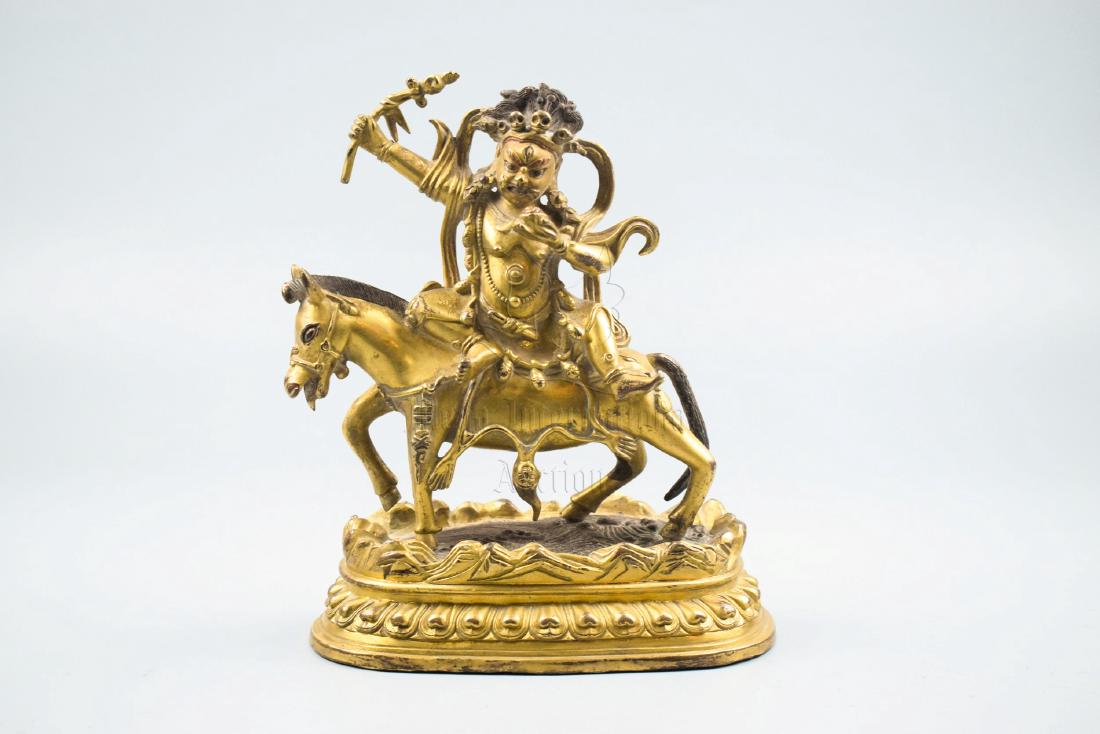GILT BRONZE CAST BODHISATTVA FIGURE: The gilt bronze Bodhisattva rides on the horse sideways on top of lotus throne pad, holding a lotus flower and raised high in one hand, wearing five-skull headdress with wrathful expression. Height: 8