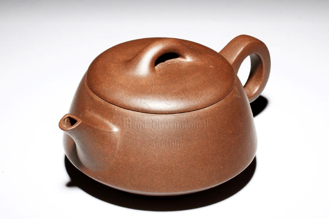 YIXING ZISHA TEAPOT: The Yixing Zisha stoneware teapot is of expanded form, the body has angular spout and handle on the sides, below a bridge shaped finial on flat lid, the clay is of dark brown color tone, and the teapo