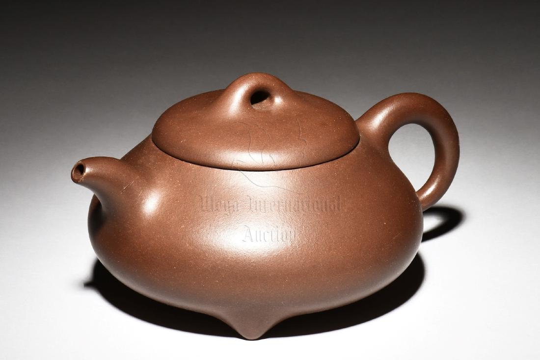 YIXING ZISHA TRIPOD TEAPOT: The Yixing Zisha stoneware teapot is of expanded form, the body stands on three short pointed feet, with curved spout and C shaped handle on the sides, and bridge shaped finial on top of flat lid, the