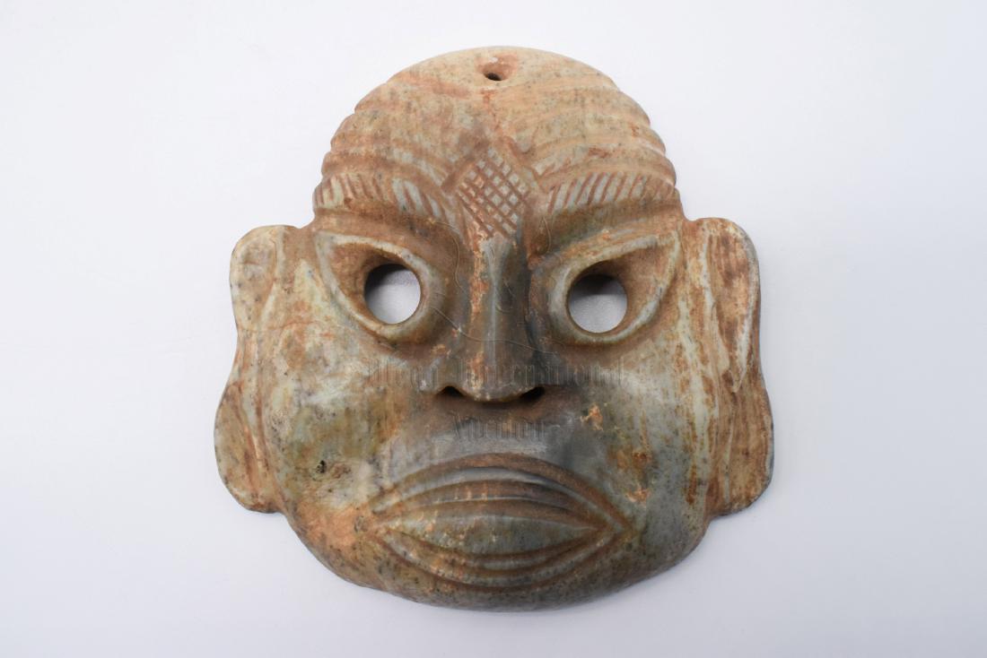 HONGSHAN CULTURE JADE CARVED 'MASK' ORNAMENT: The jade ornament carved in the form of a stylized mask, with large ears, face, and mouth, the two eyes hollow, additional stringing hole at the top of head and through the nose holes, the jade is of