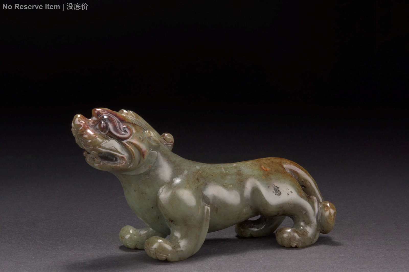 CARVED CELADON JADE PIXIU: 青玉獸 The figure depicts Pixiu standing with head raised slightly, carved from celadon jade with mottled grayish-green color and russet. CARVED CELADON JADE PIXIU Height: 4 in (10.2