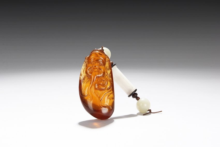 AN AMBER CARVED SHOULAO WITH WHITE JADE ORNAMENTS: The amber carving depicts a Shoulao with long beard and serene expressions, right hand holding a large coin, and carrying two more large coins on the back, the amber of alluring honey color with good 