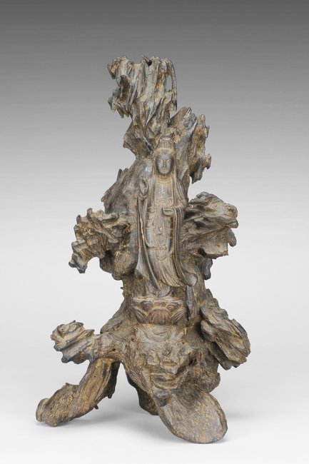 A SANDALWOOD CARVED GUANYIN FIGURE: Carved from the trunk of sandalwood, the Guanyin stands on a double lotus flowers throne, wearing long flowing robes exposing beaded ornaments on the neck, eyes closed with serene expression, against 