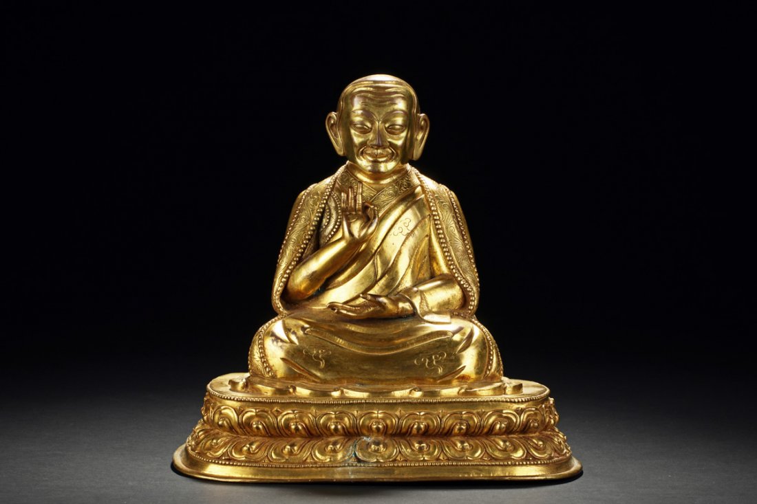 GILT BRONZE SEATED GURU: The gilt bronze guru seated on double lotus throne in dhyanasana, one hand raised at chest level with mudra, other hand rests palms up on the lap, wearing long and decorated robe, eyes open with smili