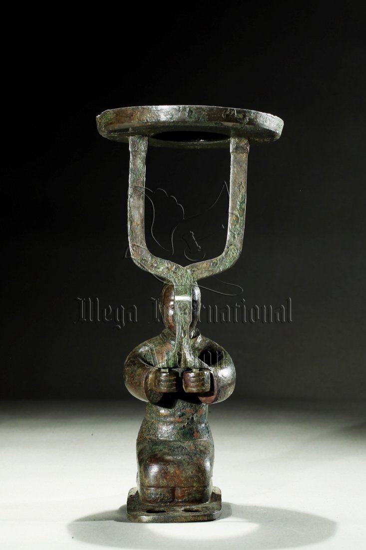 ARCHAIC BRONZE 'PERSON' LAMP: The archaic bronze lamp depicts a kneeling man with two hands held in front, holding a tall fork supporting a dish for the lamp, minor discolorations. Han Dynasty period. Height: 10 1/2 in (26.7 cm) &