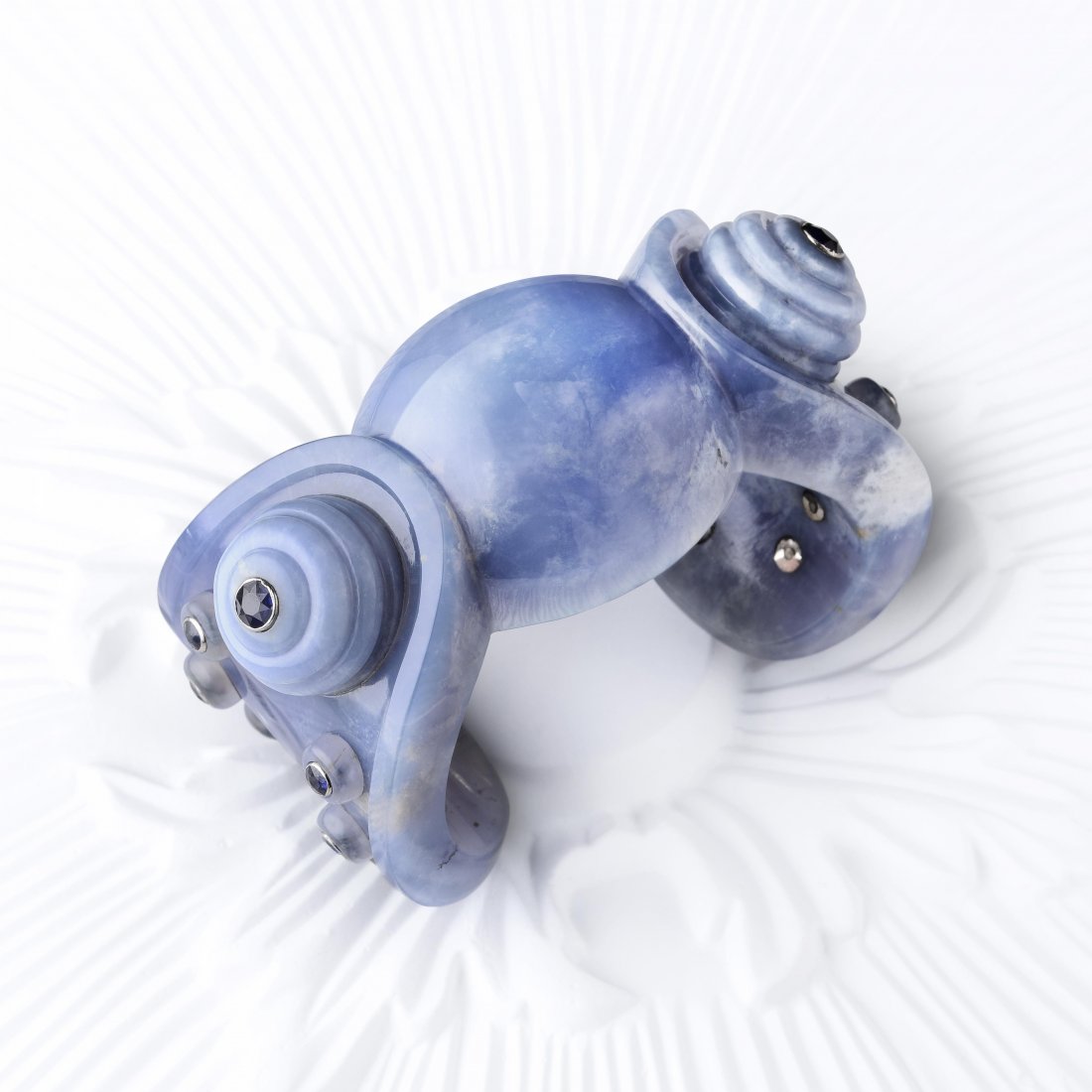 Suzanne Belperron "Cambodian" Chalcedony Cuff: A carved blue chalcedony cuff enhanced by round-cut sapphires weighing a total of approximately 3.50 carats set in gray gold; inner circumference 6 inches, breach measures 1 inch; width 1-1/2 inches;