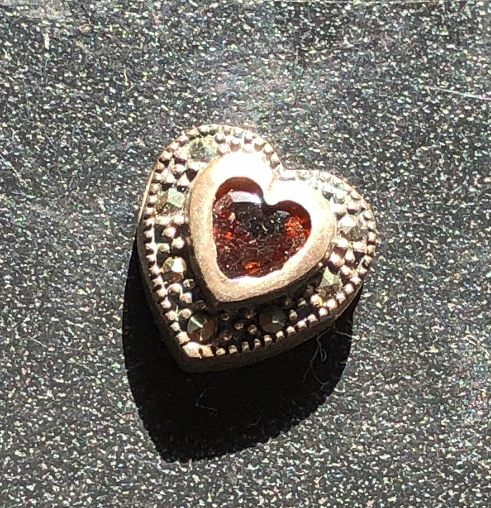 Silver Heart Pendant with Red Accent Stone: Silver heart-shaped pendant centered with a red heart-shaped stone and framed by textured beadwork, with gemstone and metal content untested.