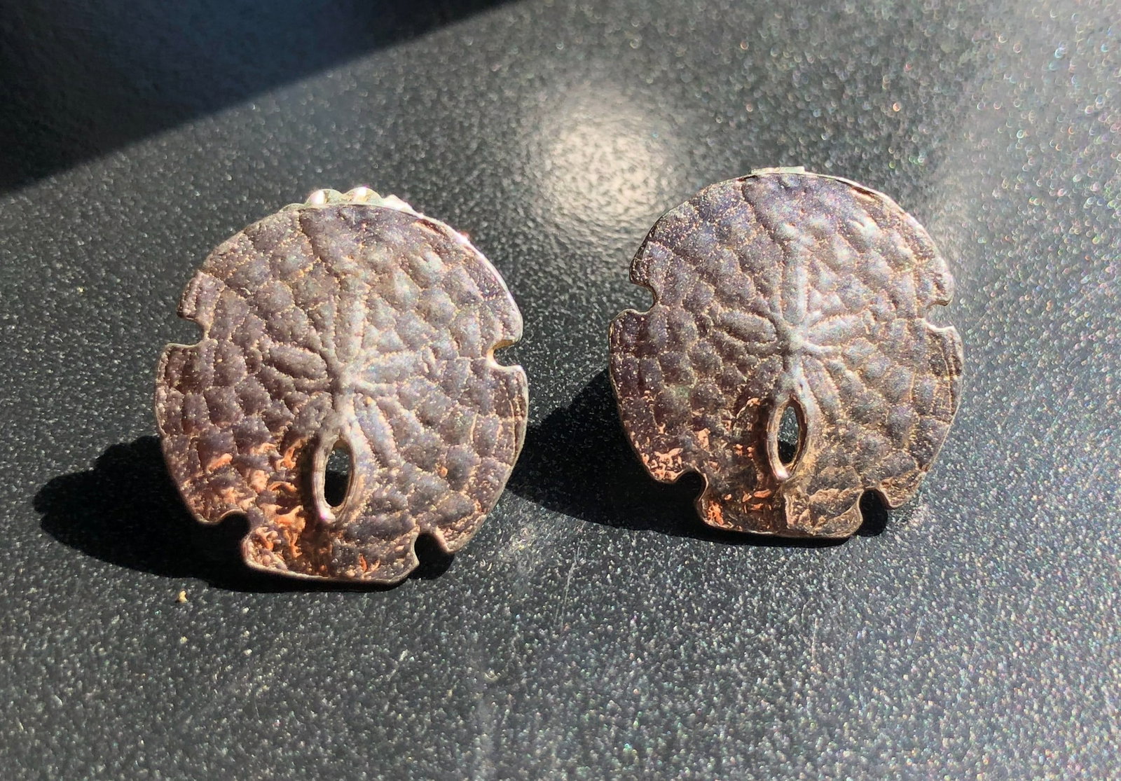 Sand Dollar Stud Silver Earrings: Pair of pierced stud Silver earrings modeled as textured sand dollars in a warm silver-to-copper tone and fitted with post backs and clear clutches, showing surface wear from previous use.
