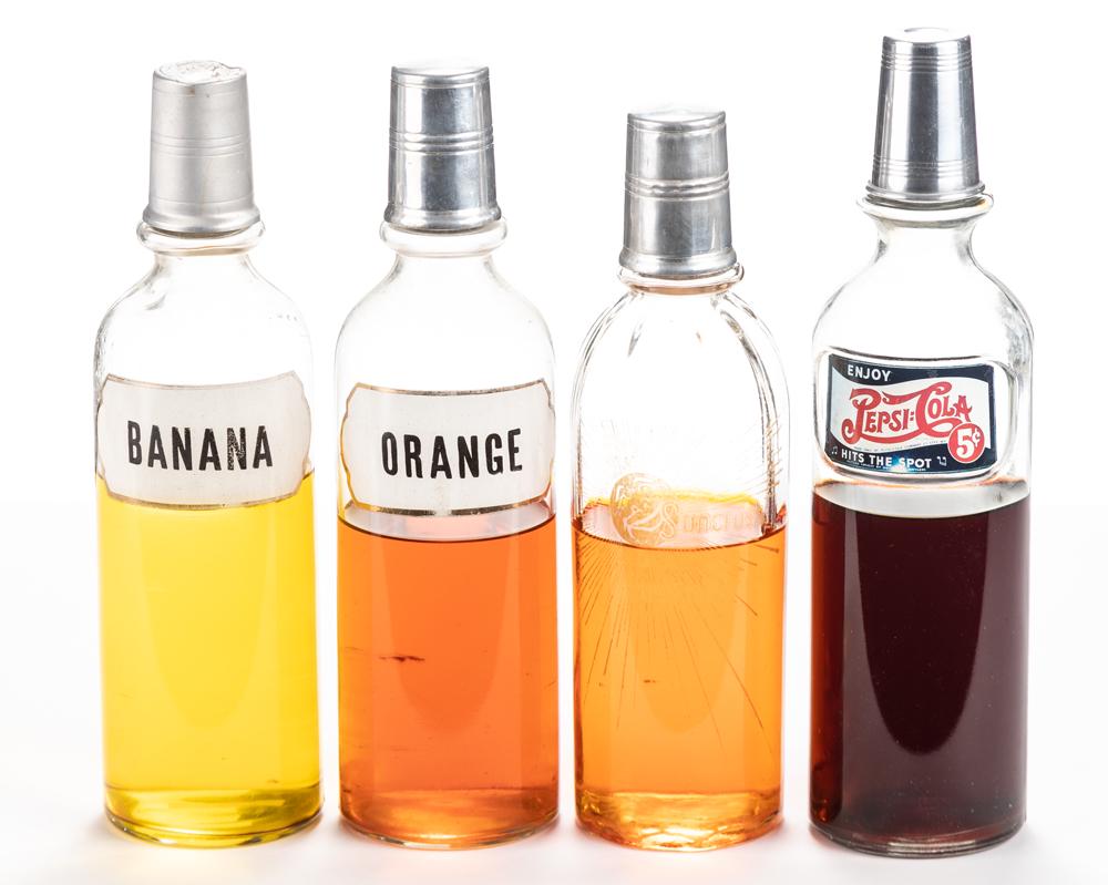 Group of four vintage glass Soda Fountain Bottles with: Group of four vintage glass Soda Fountain Bottles with original measuring cups, circa 1900. Bottles are labeled Banana / Pepsi / Sun Crush Orange / Orange. Bottles range from 11 1/2" to 12" tall. SOLD