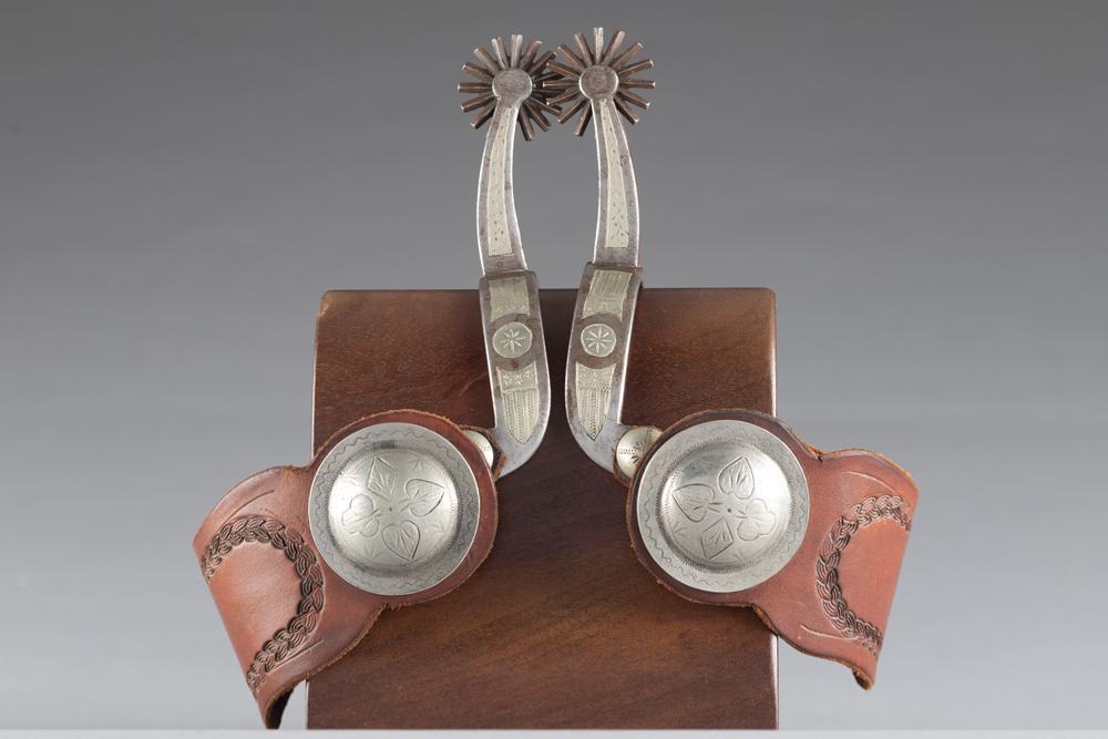 Pair of double mounted Canon City Spurs with unique drop shanks, typical prison style engraving in: Pair of double mounted Canon City Spurs with unique drop shanks, typical prison style engraving in original condition. Mounted with one-piece lightly tooled straps, 2" raised engraved conchos.