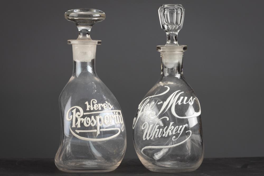 This lot consists of two early whiskey Pinch Bottles, both with ground stoppers and white enamel: This lot consists of two early whiskey Pinch Bottles, both with ground stoppers and white enamel overlay. One is marked "Fee-Mus Whiskey", 9" T. One is marked "Here's Prosperity" and measures 8 1/2" T