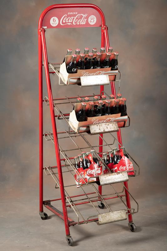 Vintage Coca-Cola Bottle Display Rack with four slanted shelves, sold with two 12 pack cartons and: Vintage Coca-Cola Bottle Display Rack with four slanted shelves, sold with two 12 pack cartons and two 6 pack cartons with unopened Coca-Cola bottles. Rack measures 60" T x 20" W x 24" D at base. Orig