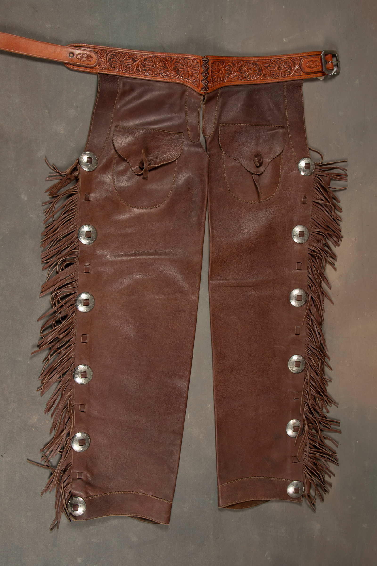 Pair of leather and fringe Stove Pipe Step In Chaps with outside sewn pockets and leather buttons,: Pair of leather and fringe Stove Pipe Step In Chaps with outside sewn pockets and leather buttons, fancy floral tooled belt marked "Miles City Saddlery Maker, Miles City, Montana". Chaps have heavy fr