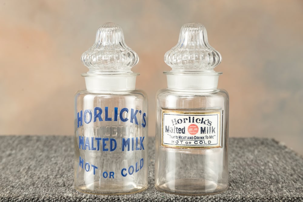 Two glass, vintage Advertising Jars with ground stoppers. One has porcelain label advertising: Two glass, vintage Advertising Jars with ground stoppers. One has porcelain label advertising "Horlick's Malted Milk / That's Meat and Drink To Me / Hot or Cold". The other has blue enamel "HORLICK'S
