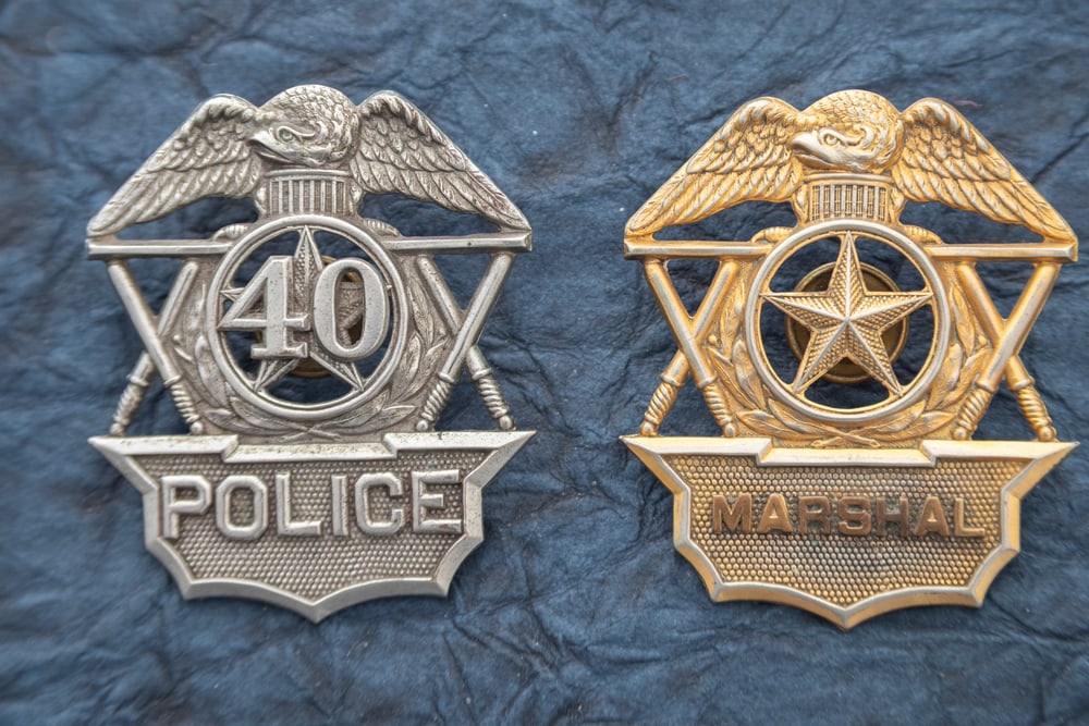 This lot consists of two Badges to include: (1) Vintage gold tone Hat Badge with Eagle Crest marked: This lot consists of two Badges to include: (1) Vintage gold tone Hat Badge with Eagle Crest marked “MARSHAL” with 5-point cut-out Star center and crossed Baton’s on each side. No markings o