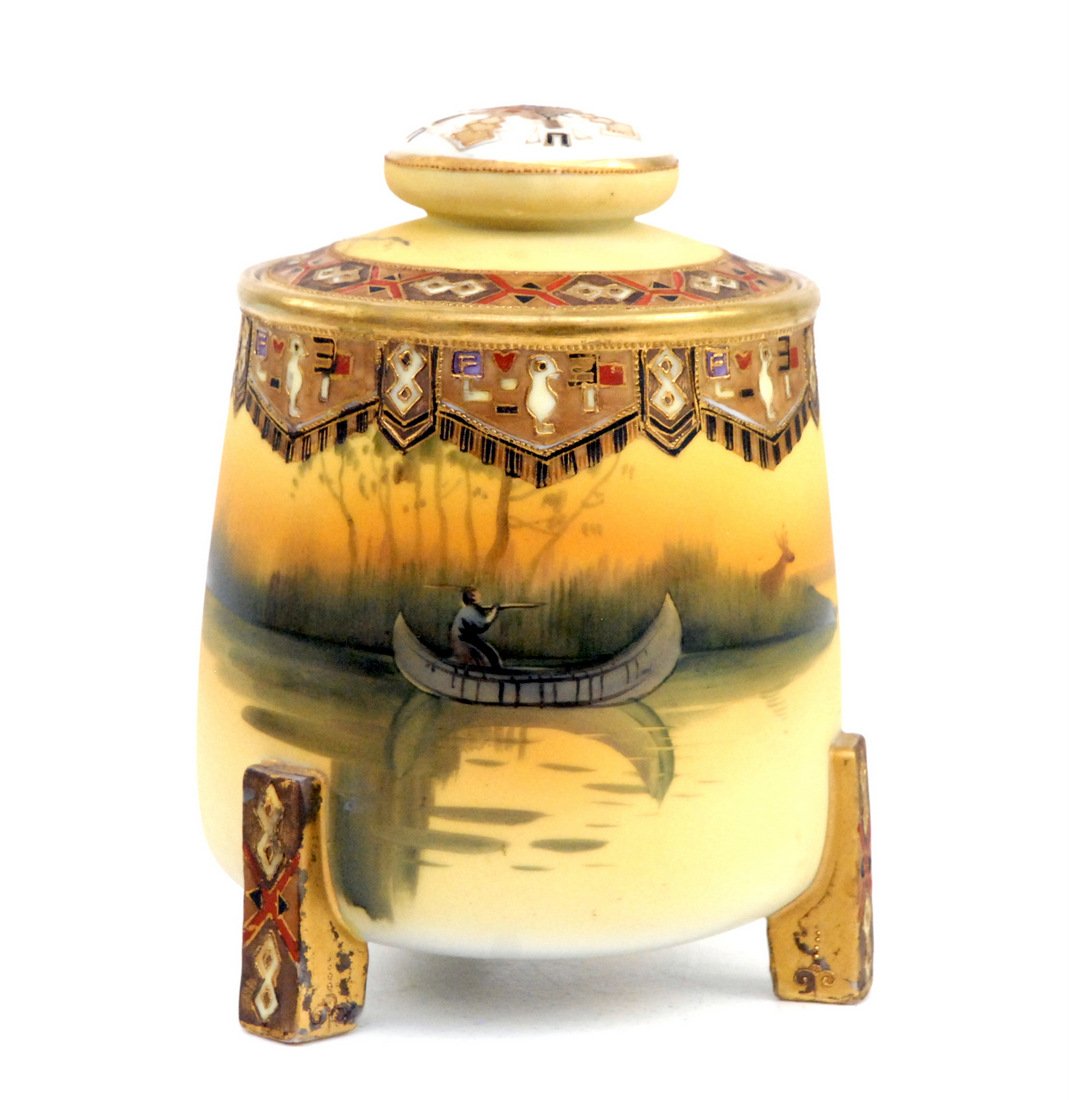 Nippon Scenic Humidor with Indian in Canoe: Tri-footed humidor with scene of Indian aiming at deer with rifle and Indian motifs in gold. 7 " high. Small touch up to one foot, otherwise fine.