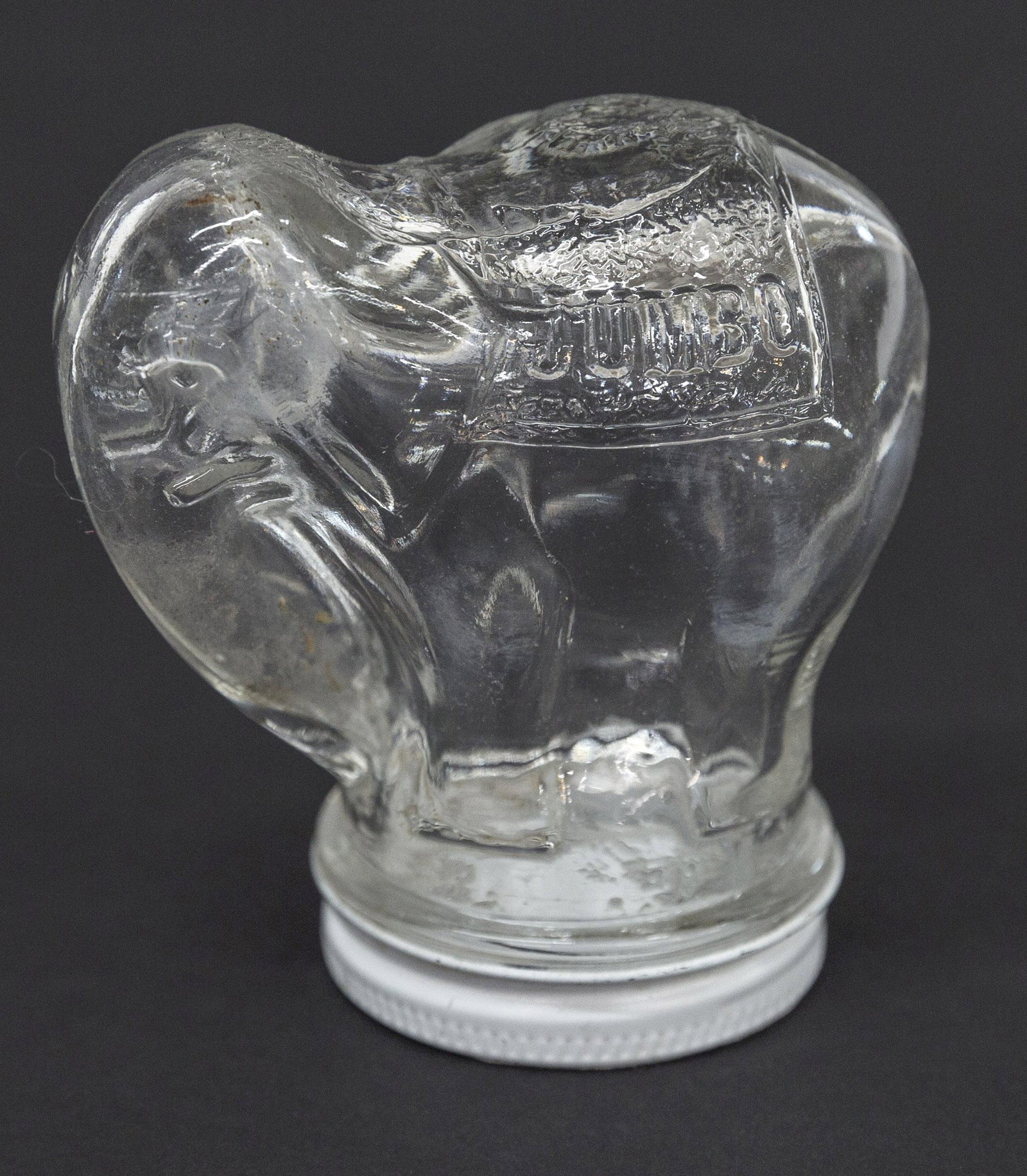 Figural Jumbo Peanut Butter Jar: Clear glass standing elephant with Jumbo on left side and Peanut Butter on right, original chromo lid with slot for bank, 3 x 1 1/2 x 3 3/8" high. Condition Fine.