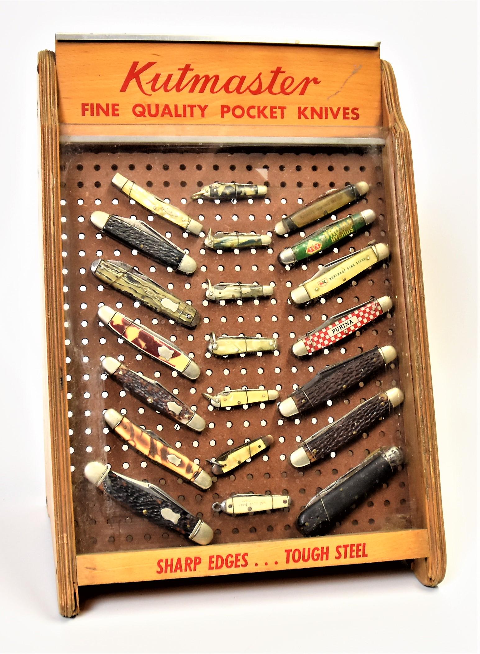 Kutmaster Pocket Knife Counter Display Case: Laminated maple with glass front and peg board back with red text reading: Kutmaster/Fine Quality Pocket Knives/Sharp Edges Tough Steel with 21 different pocket knives mounted inside, several advertis