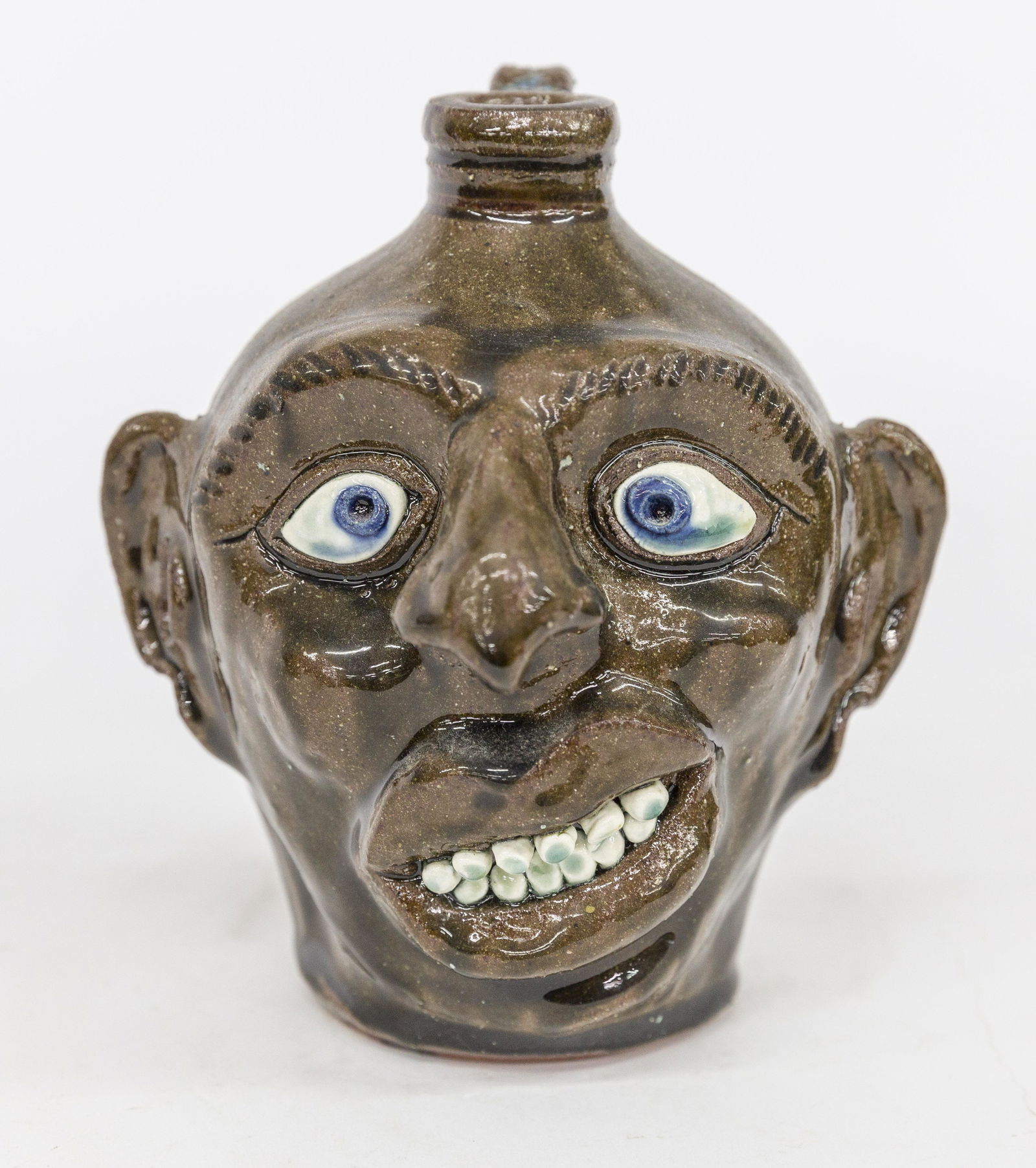 Albert Hodge Face Jug: Brown alkaline glazed redware with applied porcelain eyes and teeth, stamped on base and back Albert Hodge Vale N.C. with applied C scroll handle, 5 1/2" diameter x 7" high. Condition Fine.