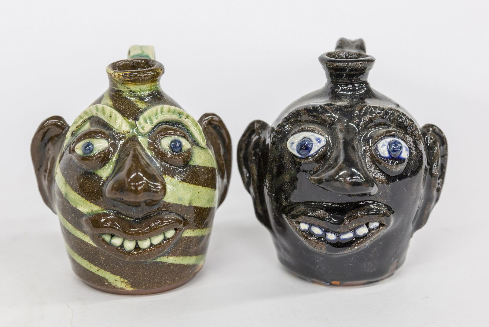 Two Albert Hodge Face Jugs: Both Alkaline glaze redware with applied porcelain eyes and teeth, C scroll handles, stamped on back and bases Albert Hodge Vale N.C., one with greenish stripes on face, each circa 4 1/2" high. Condit
